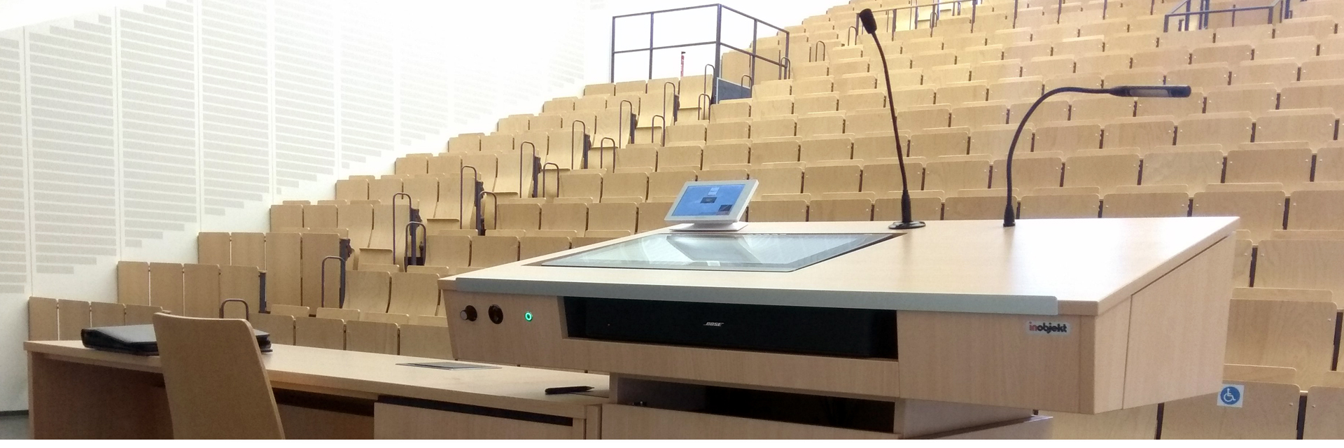 Header: Lecturer's desk teach.magna with connected lecturer's workstation in a university lecture hall. The desk contains an interactive display, media control, microphone, desk light and sound bar.